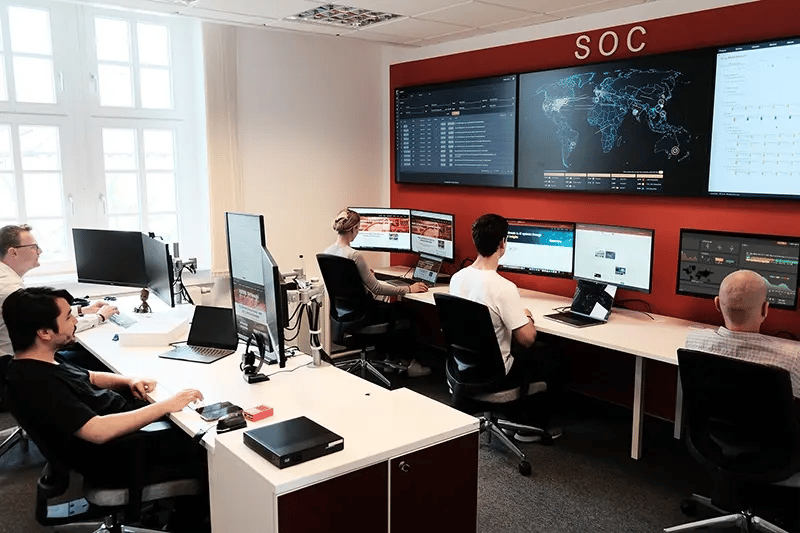 RIEDEL SOC employees sit at desks in a room with large monitors on the wall. The screens show world maps, security data and dashboards. Additional monitors and laptops on the desks.